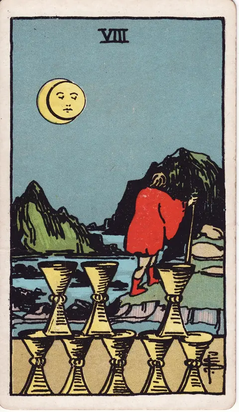 A red-cloaked traveler with a staff walks away from eight cups toward rocky mountains beneath a moonlit sky.
