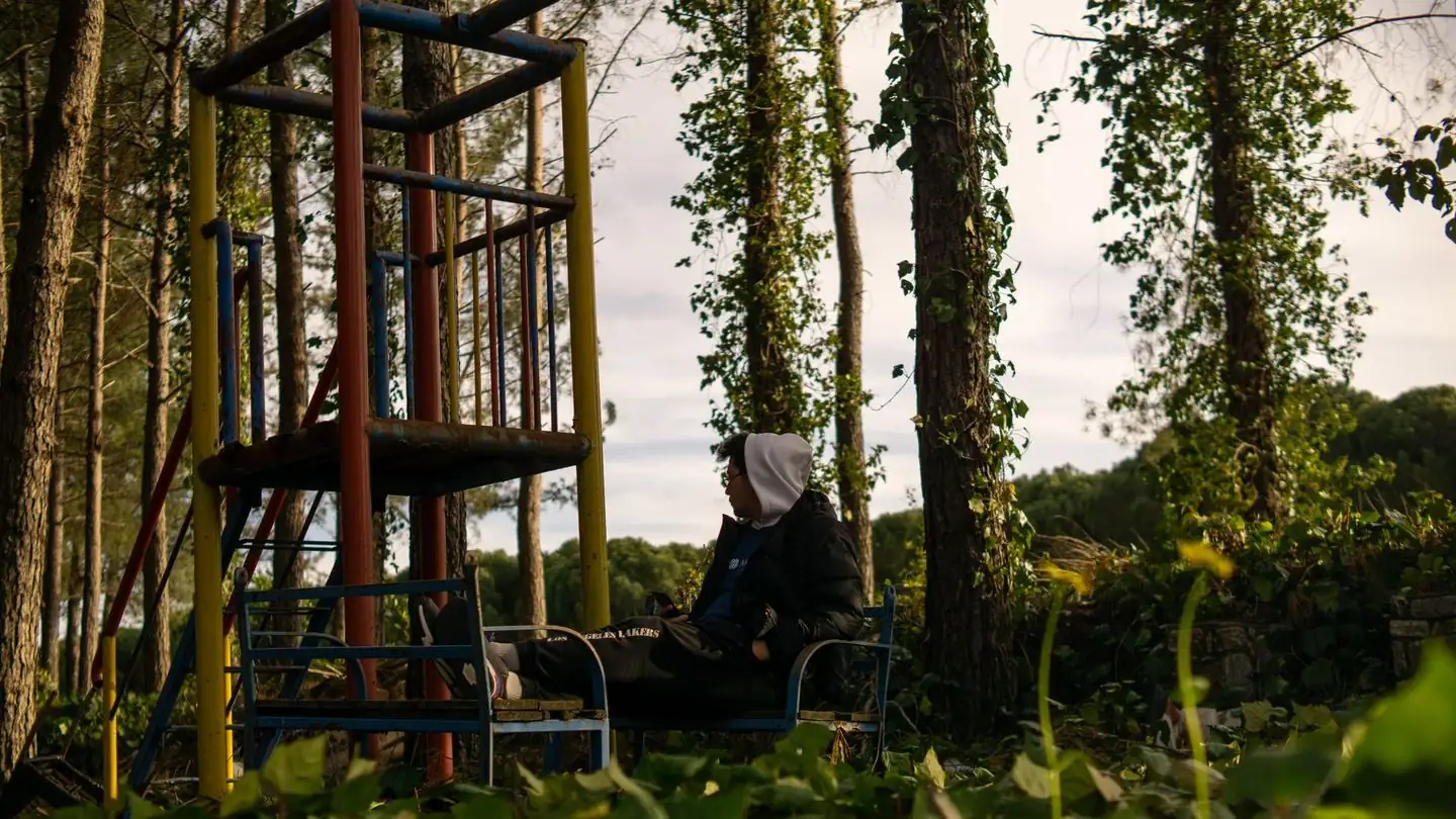 A person sits alone amid trees near a playground.