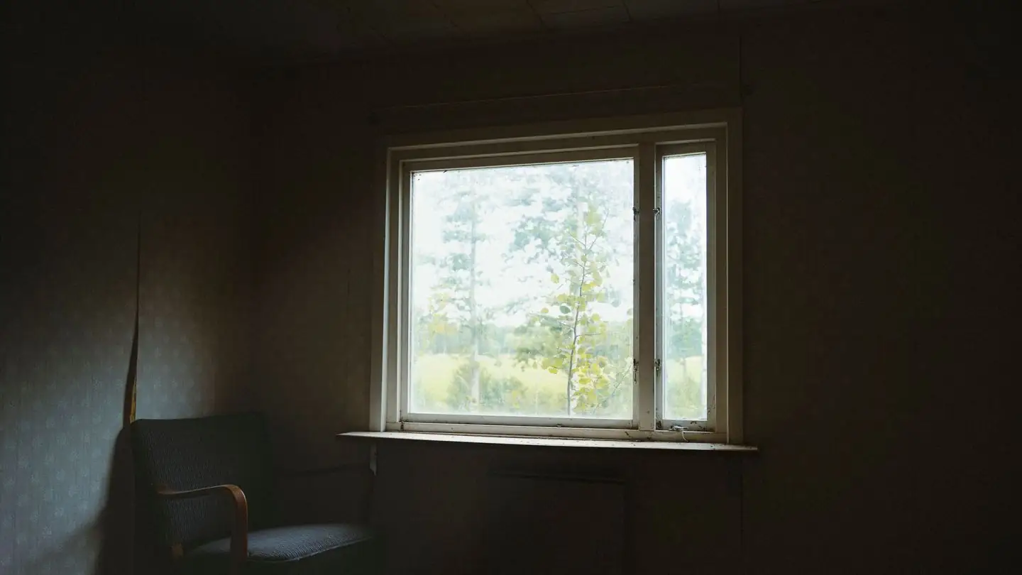 A chair stands beside a window in a dark, quiet room.