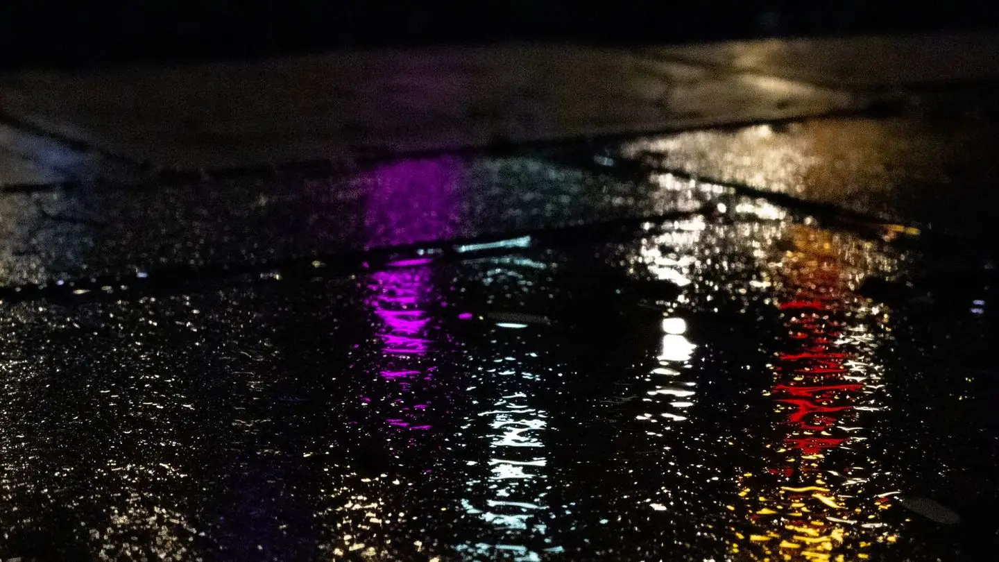 Colored neon lights reflect on wet pavement.
