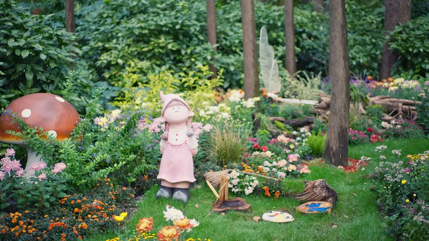 A fairy statue stands among flowers and garden ornaments.