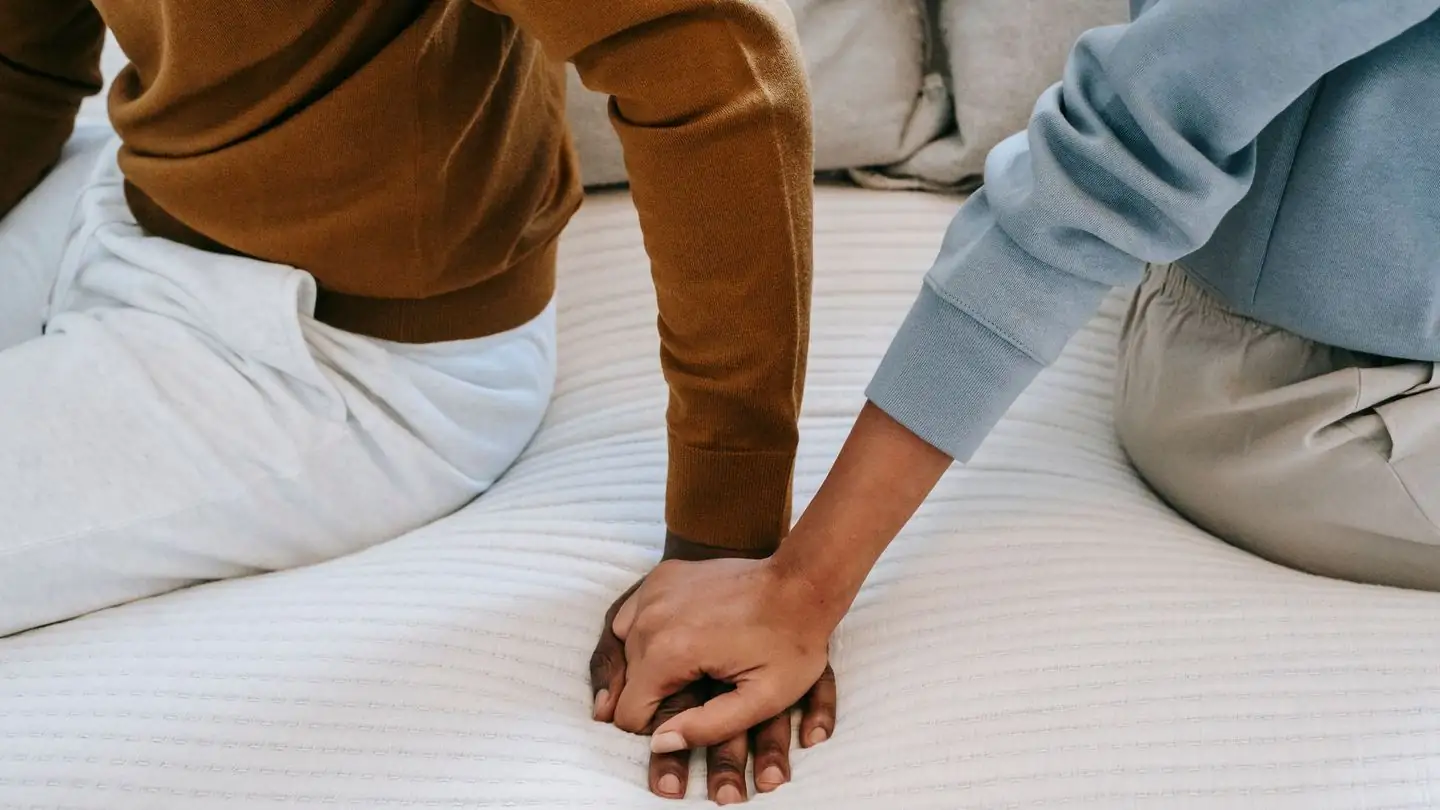 A couple sits back to back on a bed, holding hands.